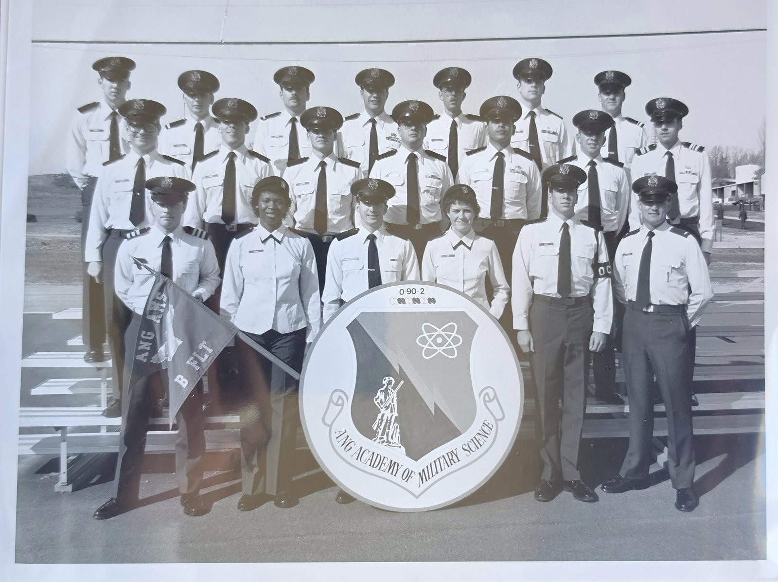 Black-and-white group photograph of about twenty cadets in service uniform standing on bleachers around a large round shield reading ANG Academy of Military Science, with the number 0-90-2 above it, and a guidon reading ANG AMS, B FLT